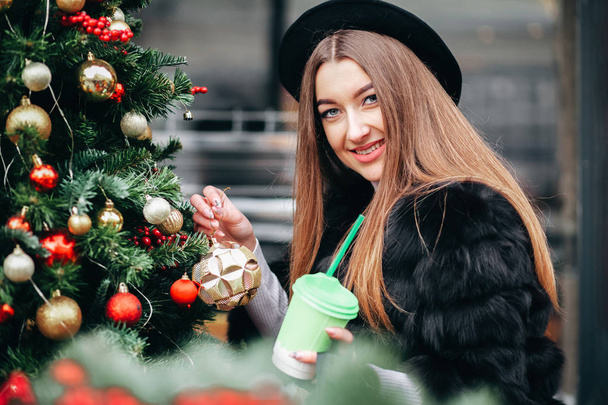 Portrait of a cute girl in a hat near Christmas decorations with a christmas ball in her hands - Fotó, kép