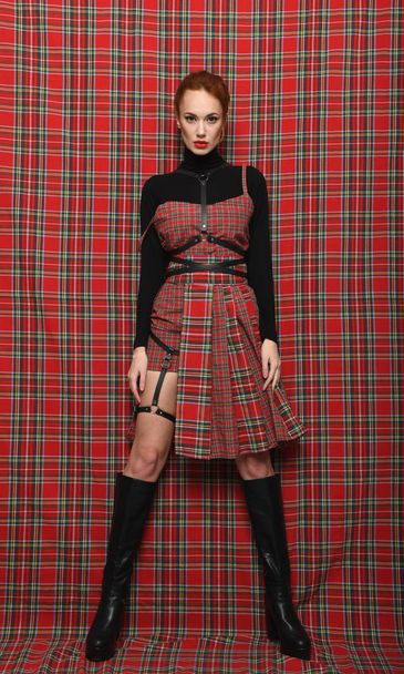 A girl in a plaid (checkered) dress on a plaid background in black golf with a bare leg demonstrates a modern stylish fashionable pose. - Photo, Image