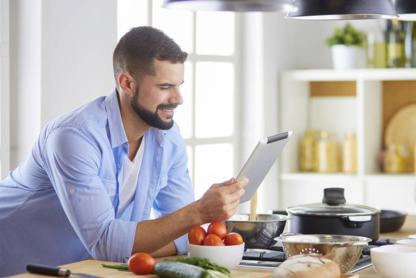 Man following recipe on digital tablet and cooking tasty and healthy food in kitchen at home - 写真・画像