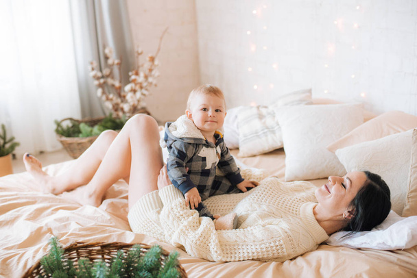 Young mother and little toddler son in a Scandinavian style bedroom decorated with Christmas garlands and needles on a large bright bed. Christmas mood - Fotoğraf, Görsel