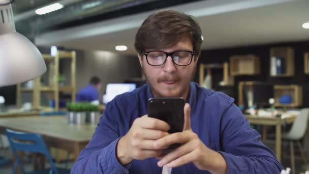 Portret Happy Young Mana Używa smartfona siedząc w biurze. Man Przegląda Internet, Ogląda filmy i korzysta z serwisów społecznościowych - Materiał filmowy, wideo