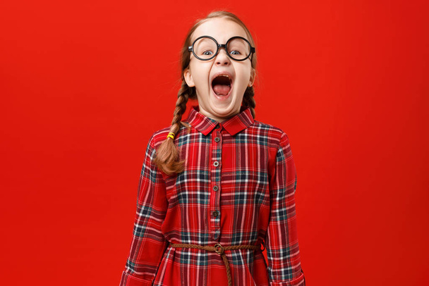 Funny little girl in glasses shouts on a red background. Portrait of a child with open mouth - Φωτογραφία, εικόνα
