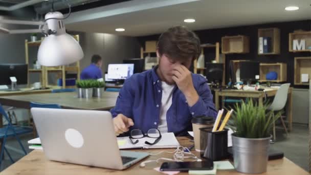 Ofis masasında dizüstü bilgisayar başında oturup kahve içmekten yorgun düşmüş genç bir adam. - Video, Çekim