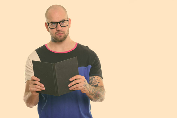 Studio shot of young bald muscular man holding book while thinking and looking up - Foto, afbeelding