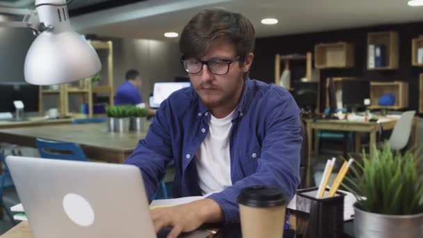 Podrážděný mladý muž s brýlemi pracující na notebooku v kanceláři - Záběry, video