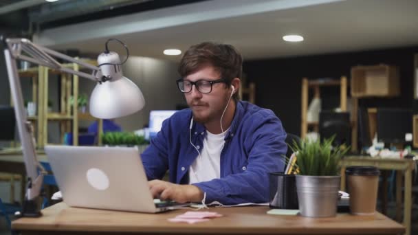 Młody programista ze słuchawkami słucha muzyki i pracuje na laptopie w biurze - Materiał filmowy, wideo