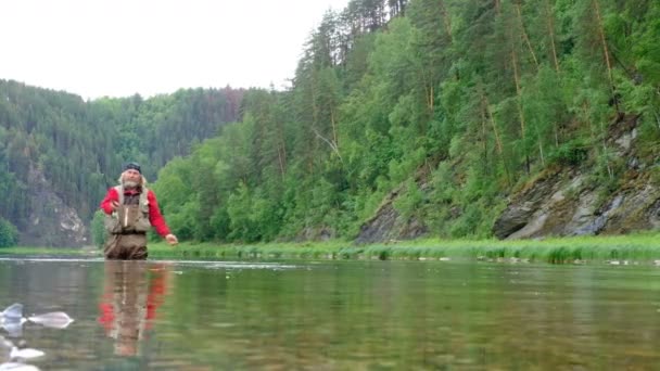 赤い服の漁師が釣竿で餌を水中に投げ込む。フライフィッシング。山の川でスポーツやエンターテイメント。全体的な計画。自然、植物、動物. - 映像、動画