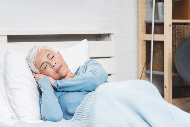 Grandma with grey hair sleep on big bed with hands under head in bright room interior  - Фото, зображення
