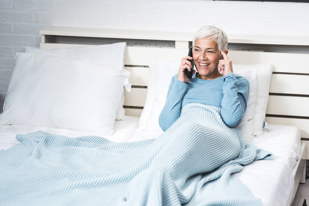 Cheerful senior woman relaxing in bed, morning awakening, talking on phone in bed still in pajamas - Fotoğraf, Görsel