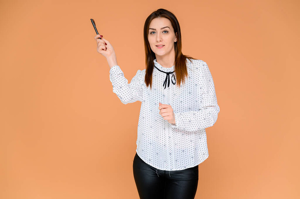 Secretary concept. Portrait of a pretty woman, manager in a white blouse with smooth dark hair. He is standing right in front of the camera, smiling, showing emotions on an orange background. - Фото, изображение
