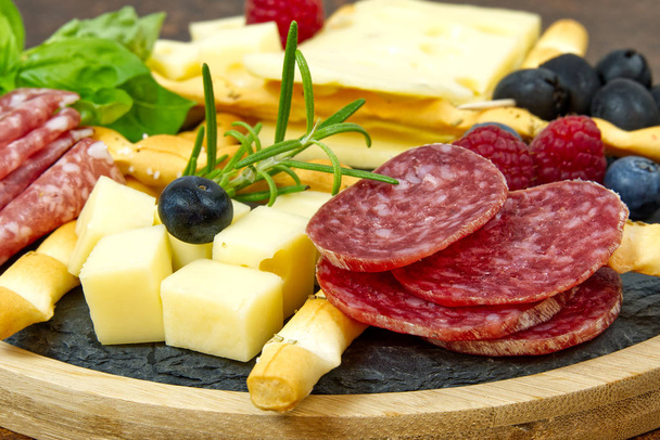 Masová a sýrová antipasti svačinka se šunkou Prosciutto, parmezánem, sýrem Blue, melounem Cantaloupe a olivami na olivovém dřevě servírovací deska na tmavém kamenném pozadí - Fotografie, Obrázek