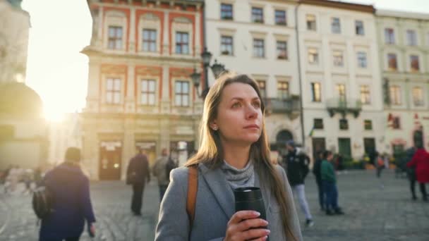 Woman with a thermos cup in hand stands in the middle of the street and looks at her watch, waiting for the one who is late to meet her at sunset on a cold autumn day - Séquence, vidéo