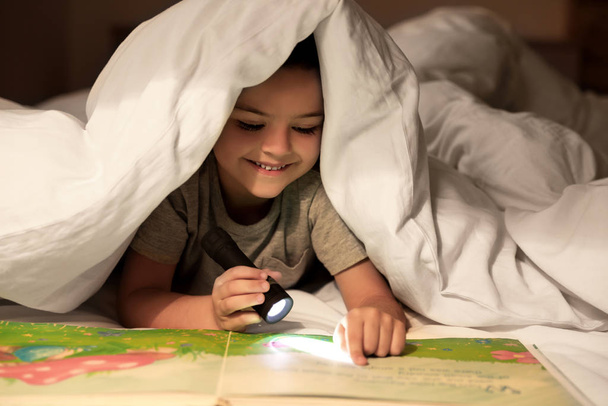 Little boy with flashlight reading book under blanket at home - Fotó, kép