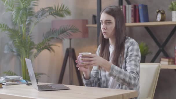 Ingerült kaukázusi lány teát vagy kávét iszik a pohárból, és gépelni kezd a laptop billentyűzetén. Dühös női szabadúszó, aki otthonról dolgozik. Távoli munka, szabadúszó, szakmai foglalkozás. - Felvétel, videó