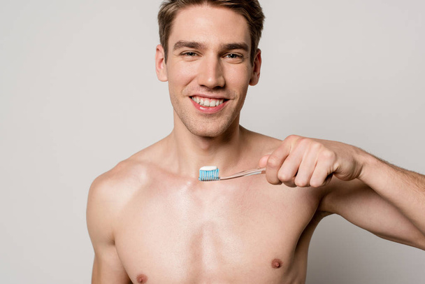 Lächeln sexy Mann mit nacktem Oberkörper hält Zahnbürste isoliert auf grau - Foto, Bild