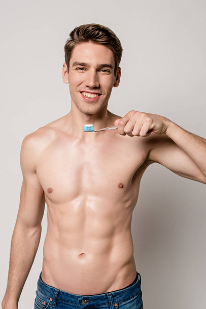 smiling sexy man with bare torso holding toothbrush isolated on grey - Fotografie, Obrázek