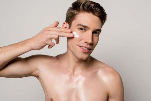 smiling sexy man with muscular torso applying face cream isolated on grey - Photo, Image