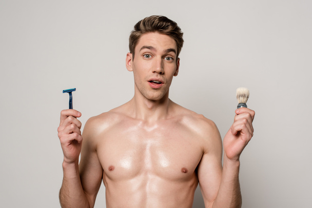 Tıraş makinesi ve tıraş fırçası tutan kaslı seksi bir adam gri renkte izole edilmiş. - Fotoğraf, Görsel
