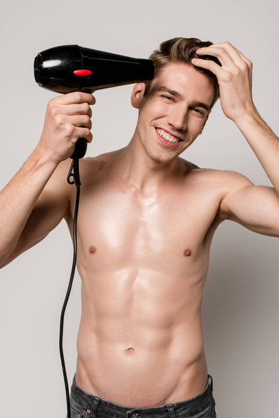 улыбающийся сексуальный мужчина с мускулистым туловищем, сушильным волосом с феном, изолированным на сером
 - Фото, изображение