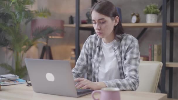 Cute Caucasian brunette girl typing on laptop, smiling and reclining on chair. Satisfied successful woman working distantly from home. Freelance, professional occupation, happiness. - Video