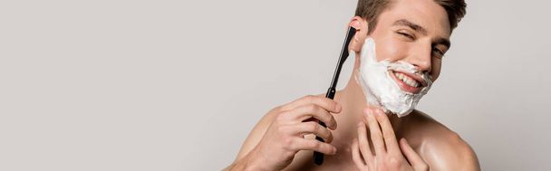 улыбающийся сексуальный мужчина с мышечным туловищем бритья с прямой бритвой изолированы на сером, панорамный снимок
 - Фото, изображение