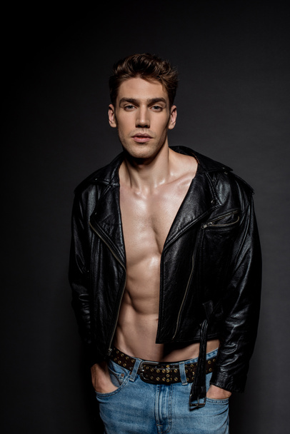 sexy young man with muscular torso in biker jacket and jeans posing with hands in pockets on black background - Photo, Image
