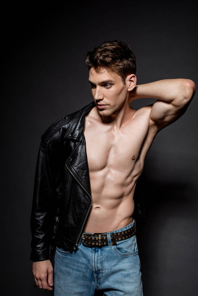 sexy young man with muscular torso putting on biker jacket and looking away on black background - Photo, Image