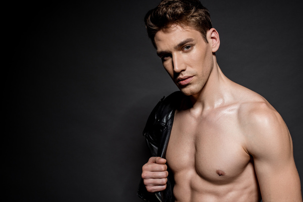 sexy young man with muscular torso and biker jacket on shoulder on black background - Photo, Image