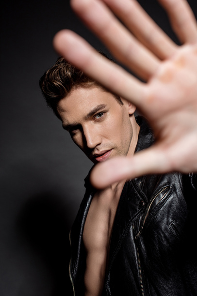 selective focus of sexy young man with muscular torso in biker jacket showing hand at camera on black background - Foto, immagini