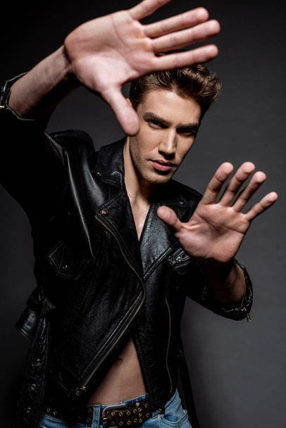 selective focus of sexy young man with muscular torso in biker jacket and jeans gesturing with hands on black background - Foto, Bild
