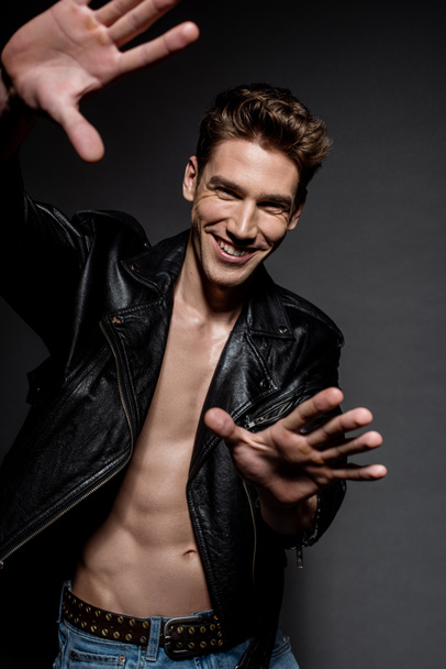 selective focus of sexy smiling man with muscular torso in biker jacket and jeans gesturing with hands on black background - Fotografie, Obrázek