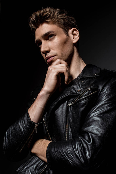 sexy young man in biker jacket touching chin and looking away on black background - Foto, afbeelding