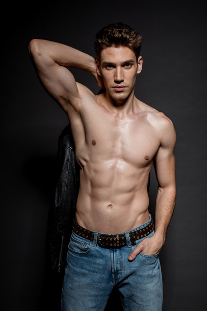 sexy young man with muscular torso and biker jacket posing with hand in pocket on black background - Foto, imagen