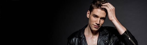 handsome sexy young man in biker jacket touching hair on black background, panoramic shot - Фото, зображення