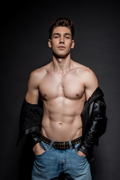 sexy young man with muscular torso in biker jacket and jeans posing with hands in pockets on black background - Photo, image