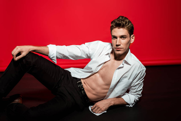 sexy young elegant man in unbuttoned shirt with muscular bare torso lying on floor on red background - Photo, image
