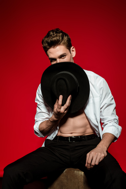 sexy young elegant man in unbuttoned shirt with muscular bare torso posing with hat near face on red background - Фото, изображение