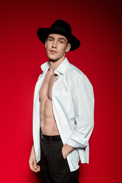 szexi fiatal elegáns férfi kigombolt ing és kalap izmos csupasz törzs pózol a piros háttér - Fotó, kép