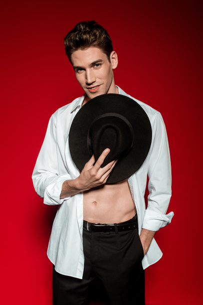 sexy young elegant man in unbuttoned shirt with muscular bare torso and hand in pocket holding hat on red background - Photo, image