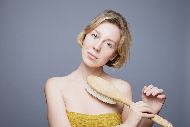 Close up portrait of European blond female, looking at camera with blue eyes, holding brush for dry massage in hand, isolated on grey studio wall background. Human face expressions, emotions concept. - Photo, Image