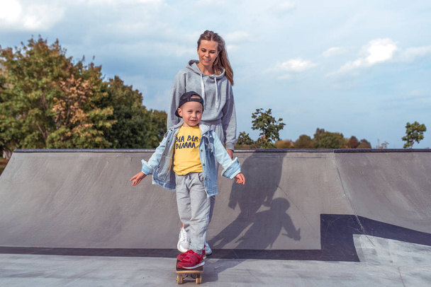 Young woman mom teaches skateboarding, little son boy, skateboard. Rest on weekend in street in park. Everyday warm clothes, autumn day. Happy smiles laugh and rejoice. Free space for copy text. - Φωτογραφία, εικόνα
