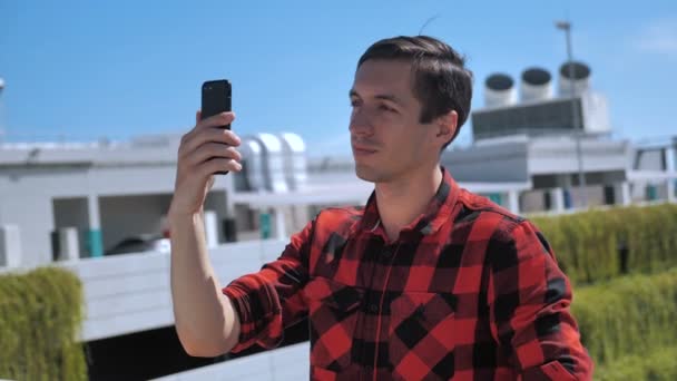 Улыбающийся молодой человек разговаривает по мобильному телефону с помощью видеосвязи на крыше. Портрет амбициозного бизнесмена, говорящего по мобильному телефону на городском фоне
.  - Кадры, видео