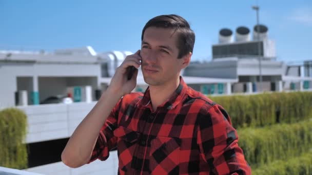 Glimlachende jonge man praat mobiele telefoon buiten op het dak. Portret van ambitieuze zakenman die buiten over mobiele telefoon praat op stedelijke achtergrond. Ondernemer bespreken van zaken via de telefoon buiten. - Video