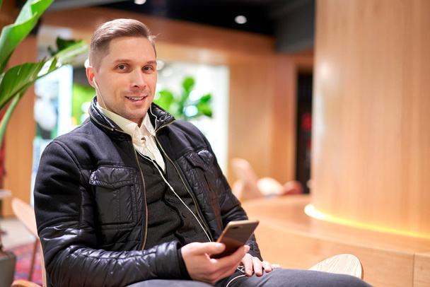 Brrunet male with smartphone sitting on chair in store against green plant - Zdjęcie, obraz