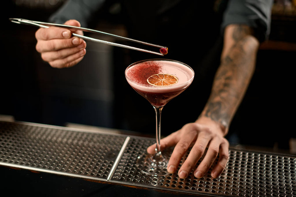 Barman versiert een alcoholische cocktail in het glas met een gedroogde sinaasappel en rode specerijen met pincet met rozenknop - Foto, afbeelding