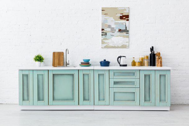 weiße und türkisfarbene Kücheneinrichtung mit Geschirr und abstrakter Malerei an der Ziegelwand - Foto, Bild