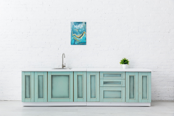 białe i turkusowe wnętrze kuchni z umywalką, rośliną i abstrakcyjnym malowaniem na ścianie z cegły - Zdjęcie, obraz