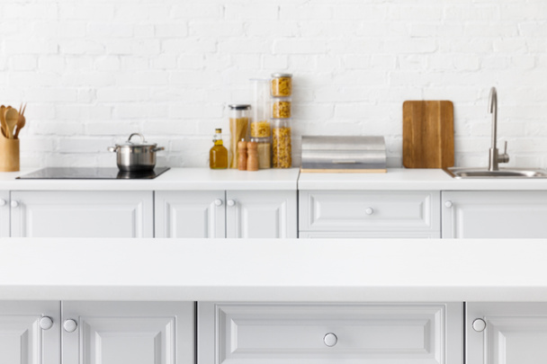 selectieve focus van lege tafelblad en minimalistische moderne witte keuken interieur met keukengerei en voedsel in de buurt van baksteen muur op de achtergrond - Foto, afbeelding