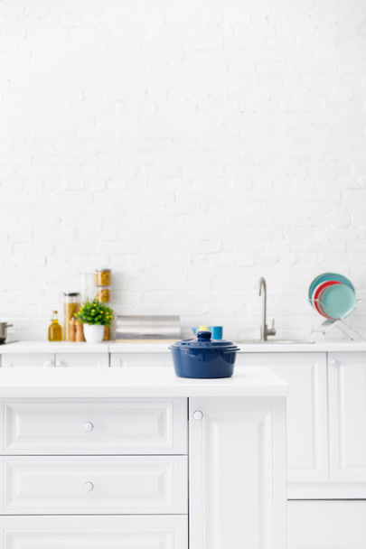 Mavi tencerenin seçici odak noktası ve arka planda mutfak eşyaları ve tuğla duvarın yanında yiyecek olan minimalist modern beyaz mutfak. - Fotoğraf, Görsel