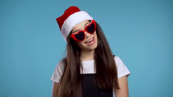 Atraktivní dívka v Santa klobouk a srdce ve tvaru slunečních brýlí slaví Vánoce nebo Nový rok na modrém pozadí. Šťastná hezká žena se usmívá a baví. - Záběry, video
